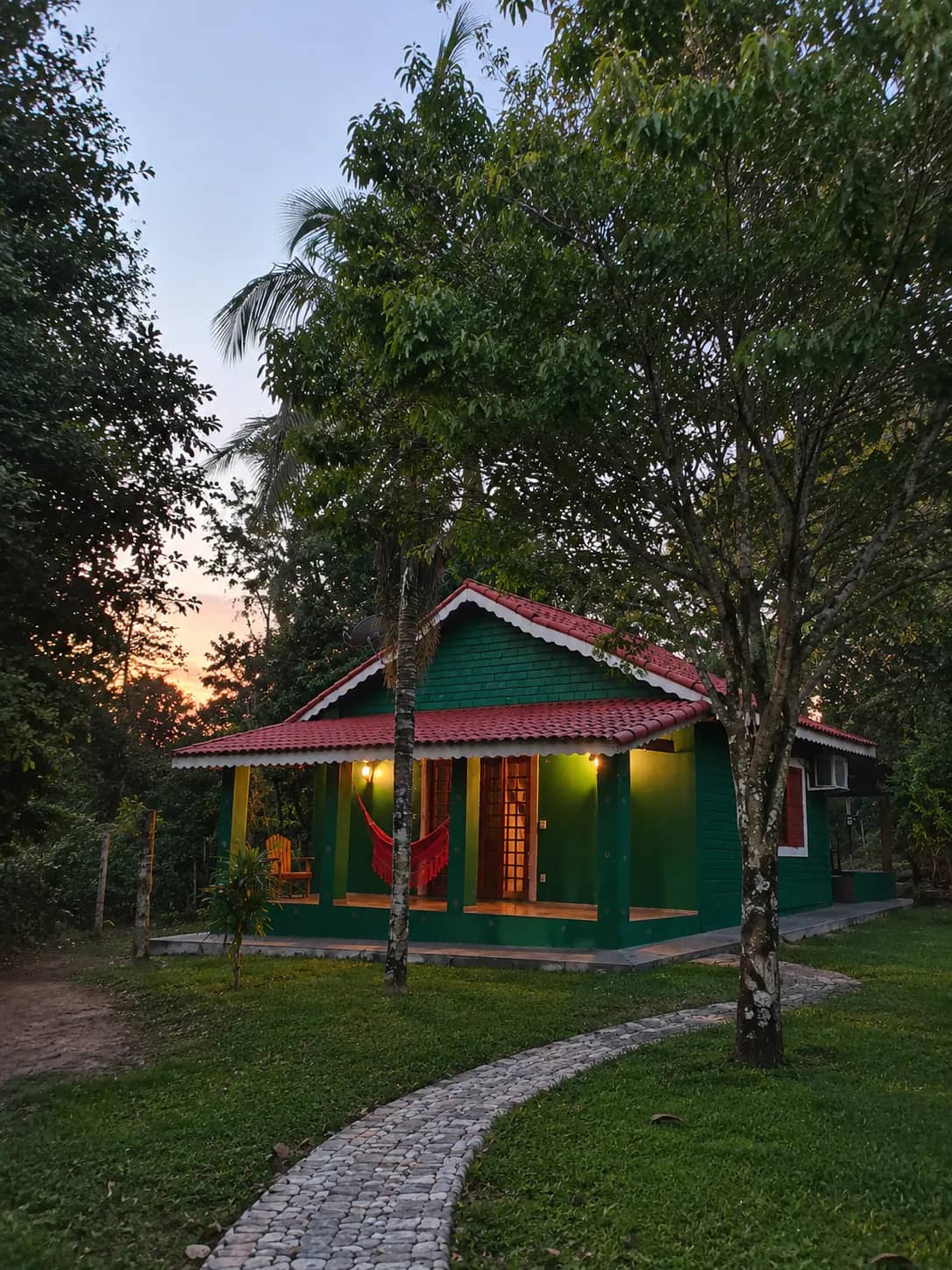 Chalé verde com varanda, rede e luzes acesas ao entardecer, cercado de árvores