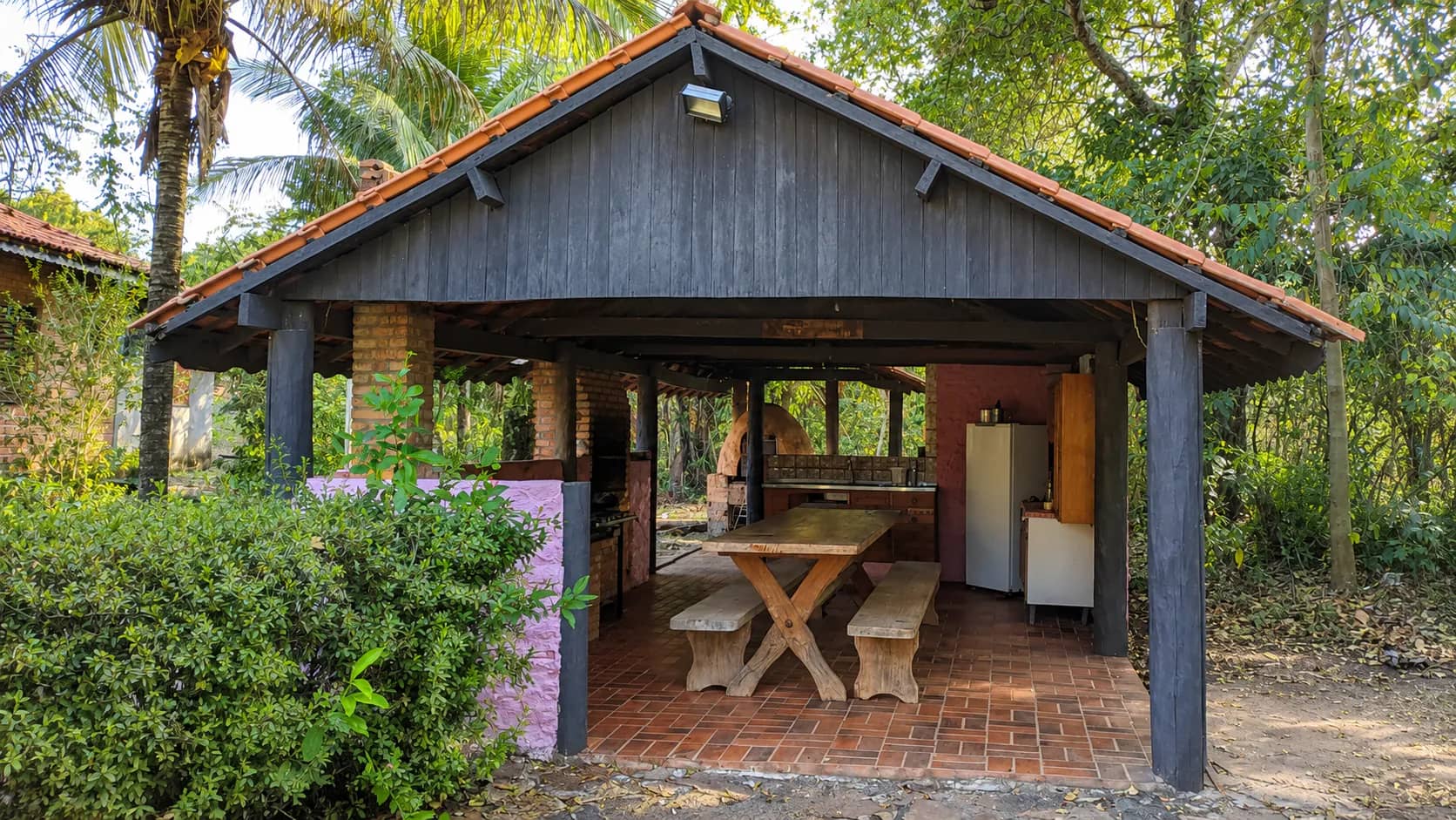 Cozinha pantaneira coberta em meio à mata, com mesa de madeira e bancos, forno de barro, churrasqueira de tijolo, pia e geladeira